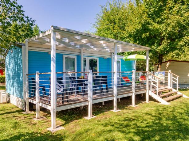 Blaues Mobilheim mit Veranda, Stühlen und grünem Sonnenschirm.