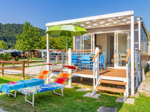 Modernes Bungalow mit Veranda, Liegestühlen und Sonnenschirm auf einem grünen Campingplatz.