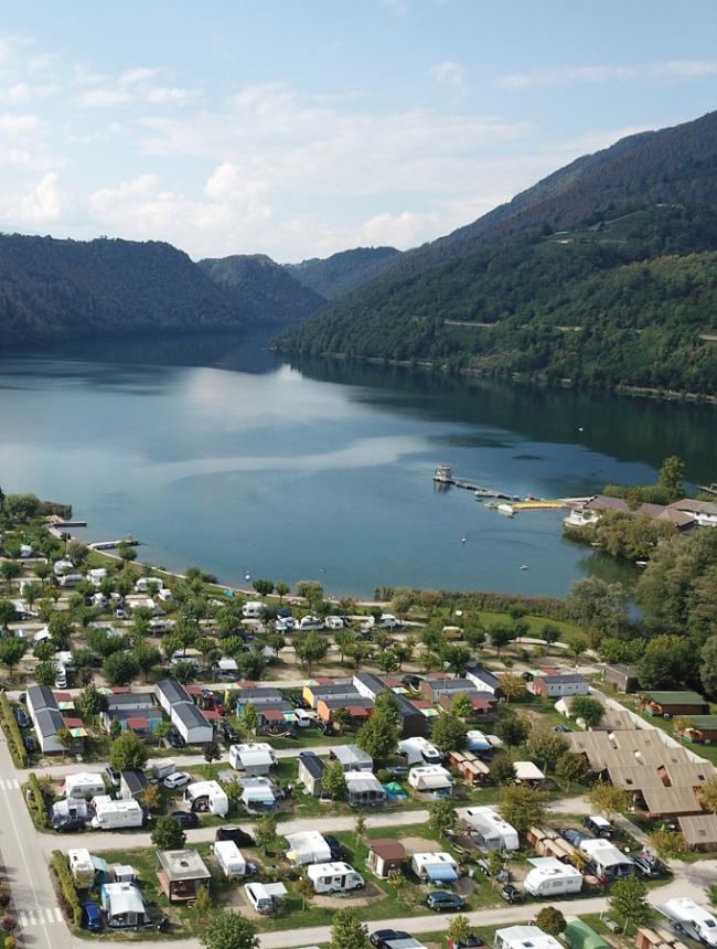 Malersiche Aussicht auf einen See mit Campingplatz und Bergen im Hintergrund.